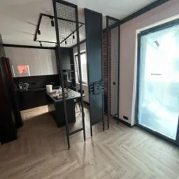 Loftowy design wnętrza z metalowymi elementami: ażurowe ścianki działowe z czarnej siatki i płaskie panele oddzielają kuchnię od okna. Parkiet w jodełkę.