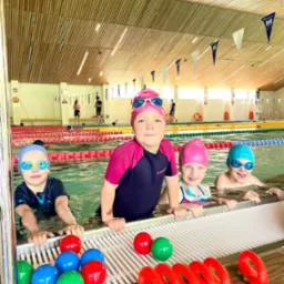 Grupa dzieci w czepkach i okularkach pływackich uśmiecha się w basenie, z kolorowymi zabawkami na brzegu. W tle widoczni inni pływacy i elementy wyposażenia basenu.