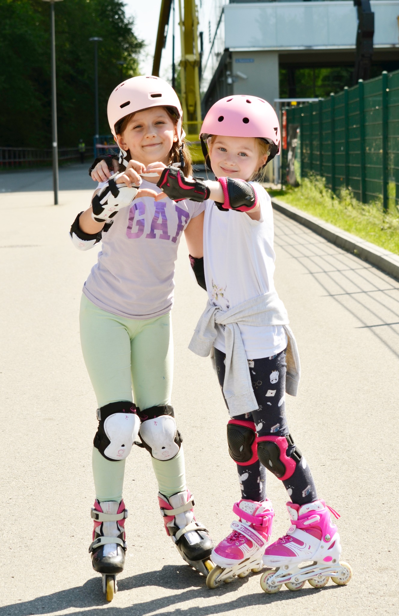 Dwie uśmiechnięte dziewczynki w kaskach i ochraniaczach, na rolkach, stoją obok siebie na chodniku, pokazując serce z dłoni na tle zieleni miejskiej.