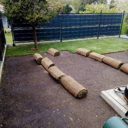 Andrii Ronskyi - Rozwinięte rolki trawy na czarnej siatce w ogrodzie z ciemnym ogrodzeniem. Częściowo ułożona trawa z rolki w ogrodzie.