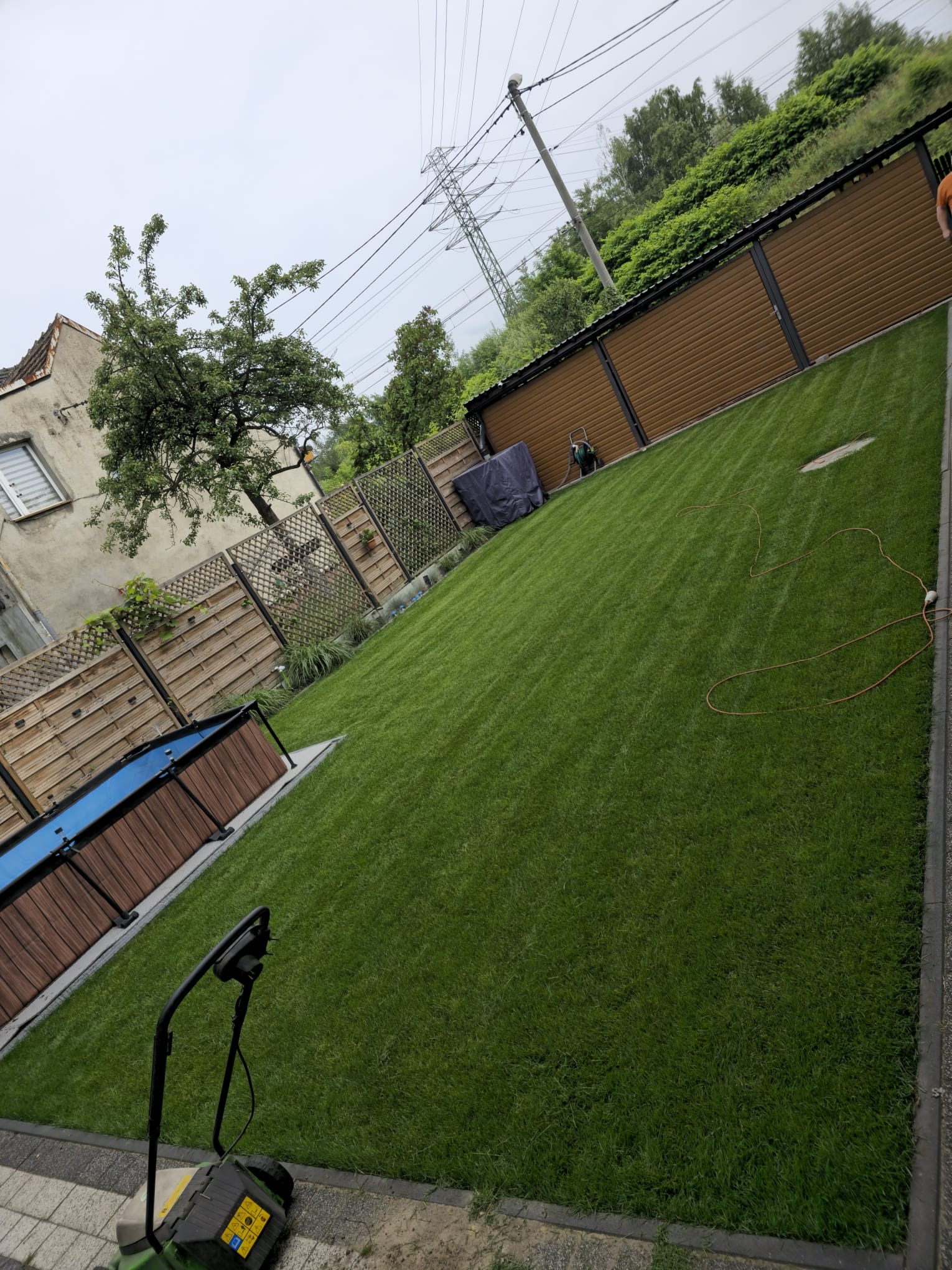 Świeżo założony trawnik z rolki, widok z góry, obok kosiarka. W tle ogrodzenie panelowe, drzewa i słup wysokiego napięcia. Perfekcyjnie równa, zielona powierzchnia.