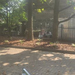 USŁUGI OGRODNICZE MICHAŁ WALA - Fragment świeżo założonego ogrodu z korą, roślinami i drzewami, widok z perspektywy wózka z kamieniami, ogrodzenie w tle, słoneczny dzień.