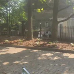 Fragment świeżo założonego ogrodu z korą, roślinami i drzewami, widok z perspektywy wózka z kamieniami, ogrodzenie w tle, słoneczny dzień.