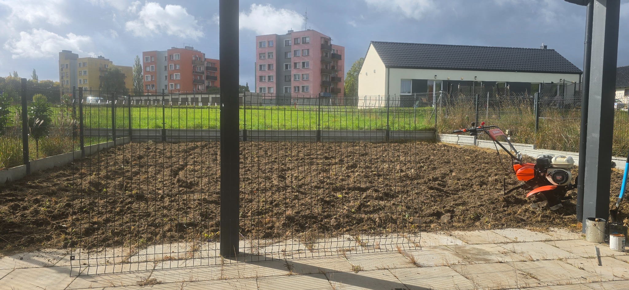 Przygotowana gleba pod trawnik w ogrodzie miejskim, z widocznym glebogryzarką i budynkami w tle. Grządka otoczona niskim, czarnym ogrodzeniem.