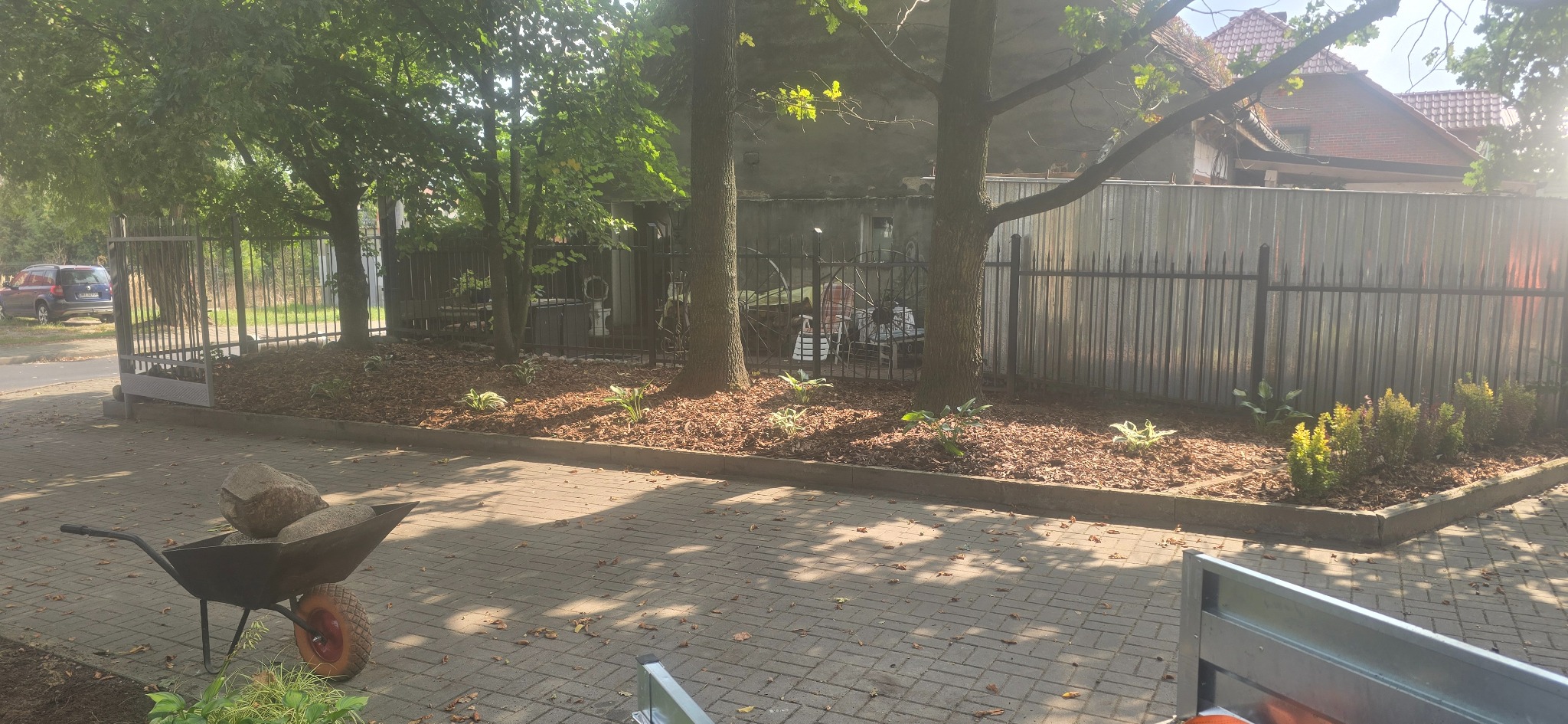 Fragment świeżo założonego ogrodu z korą, roślinami i drzewami, widok z perspektywy wózka z kamieniami, ogrodzenie w tle, słoneczny dzień.