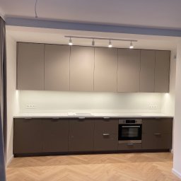 MEBLE+ - Nowoczesna kuchnia w odcieniach beżu i brązu z oświetleniem punktowym, jasnym blatem i drewnianą podłogą ułożoną w jodełkę. Widoczne zasłony po lewej stronie.