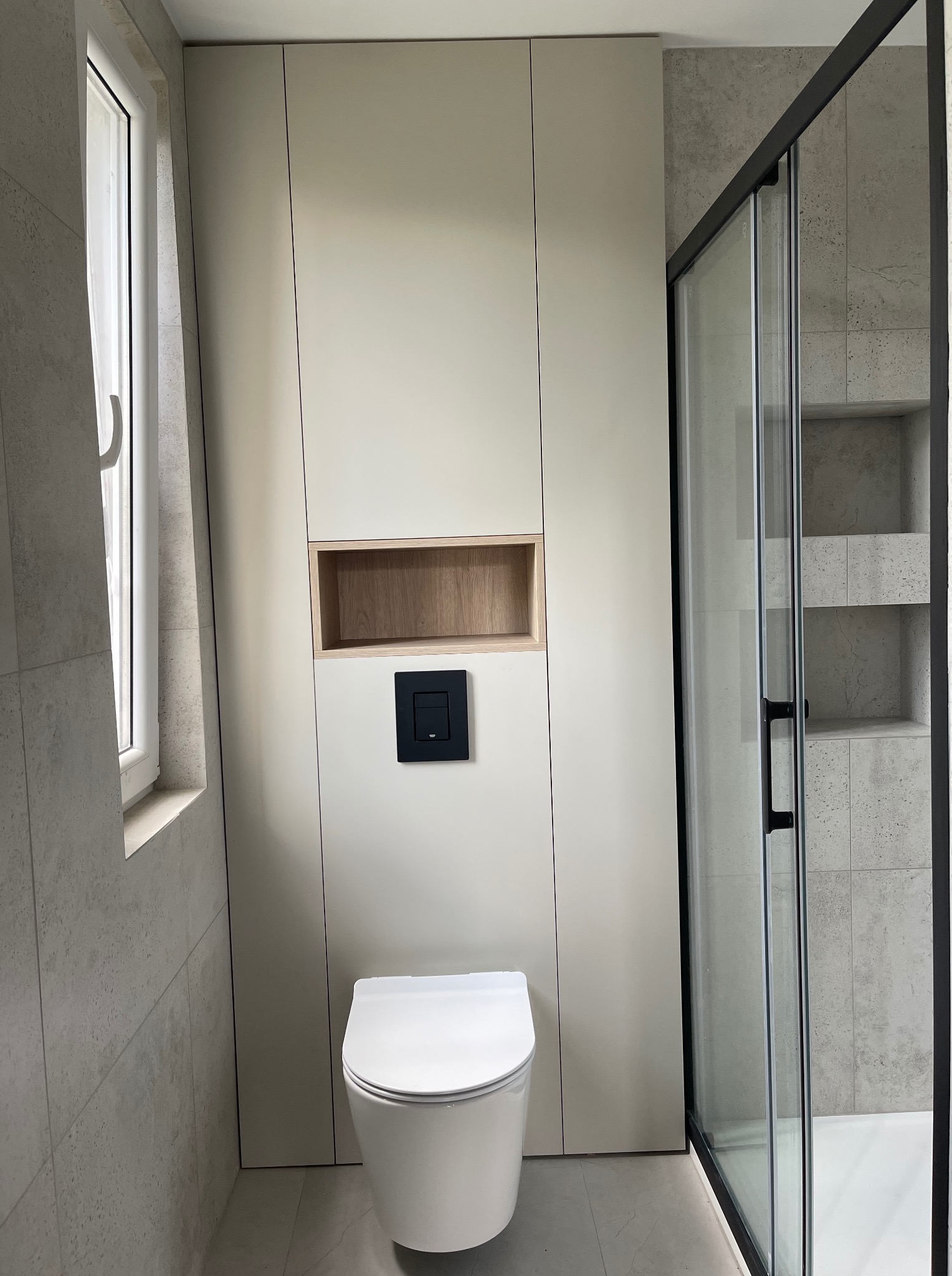 Nowoczesna łazienka z zabudową meblową w odcieniach beżu, z wnęką z drewnianym akcentem nad toaletą i kabiną prysznicową z czarnymi profilami.