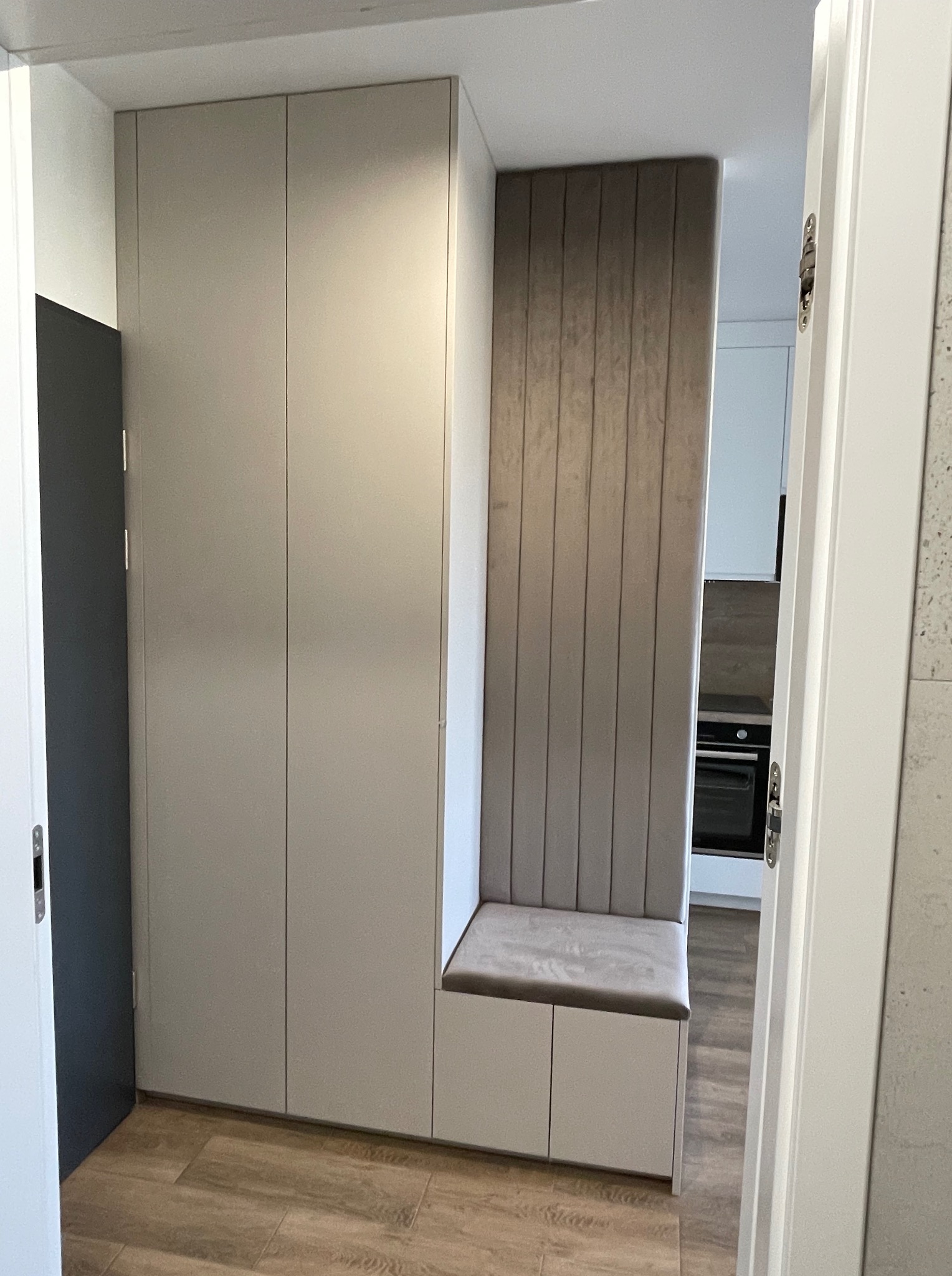 Szafa w przedpokoju z siedziskiem i tapicerowanym panelem ściennym w odcieniach beżu. Widok na kuchnię w tle. Drewnopodobna podłoga.