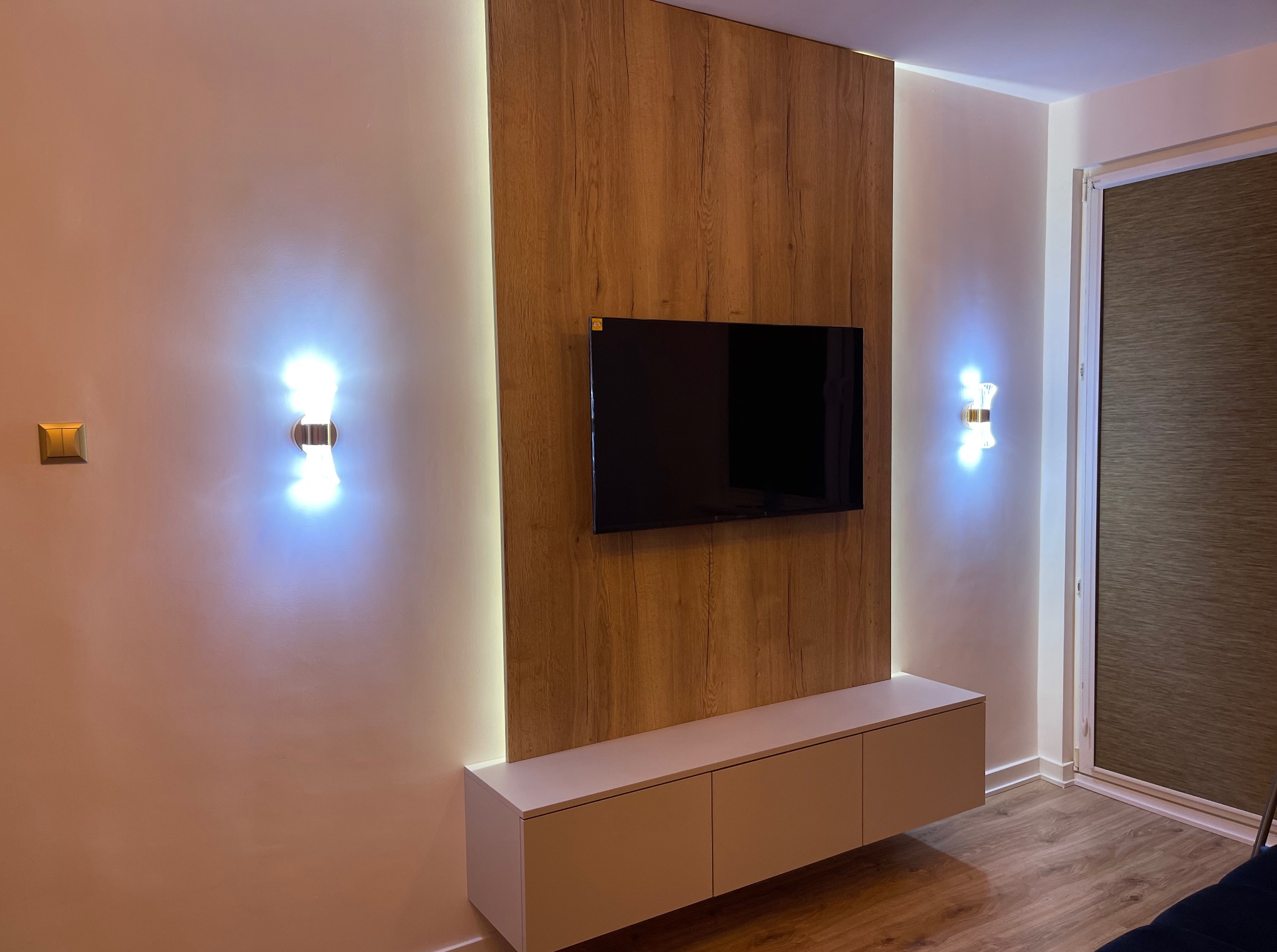 Nowoczesna zabudowa ściany z panelem drewnopodobnym, podświetleniem LED i szafką RTV. Dwa kinkiety po bokach dodają klimatu.