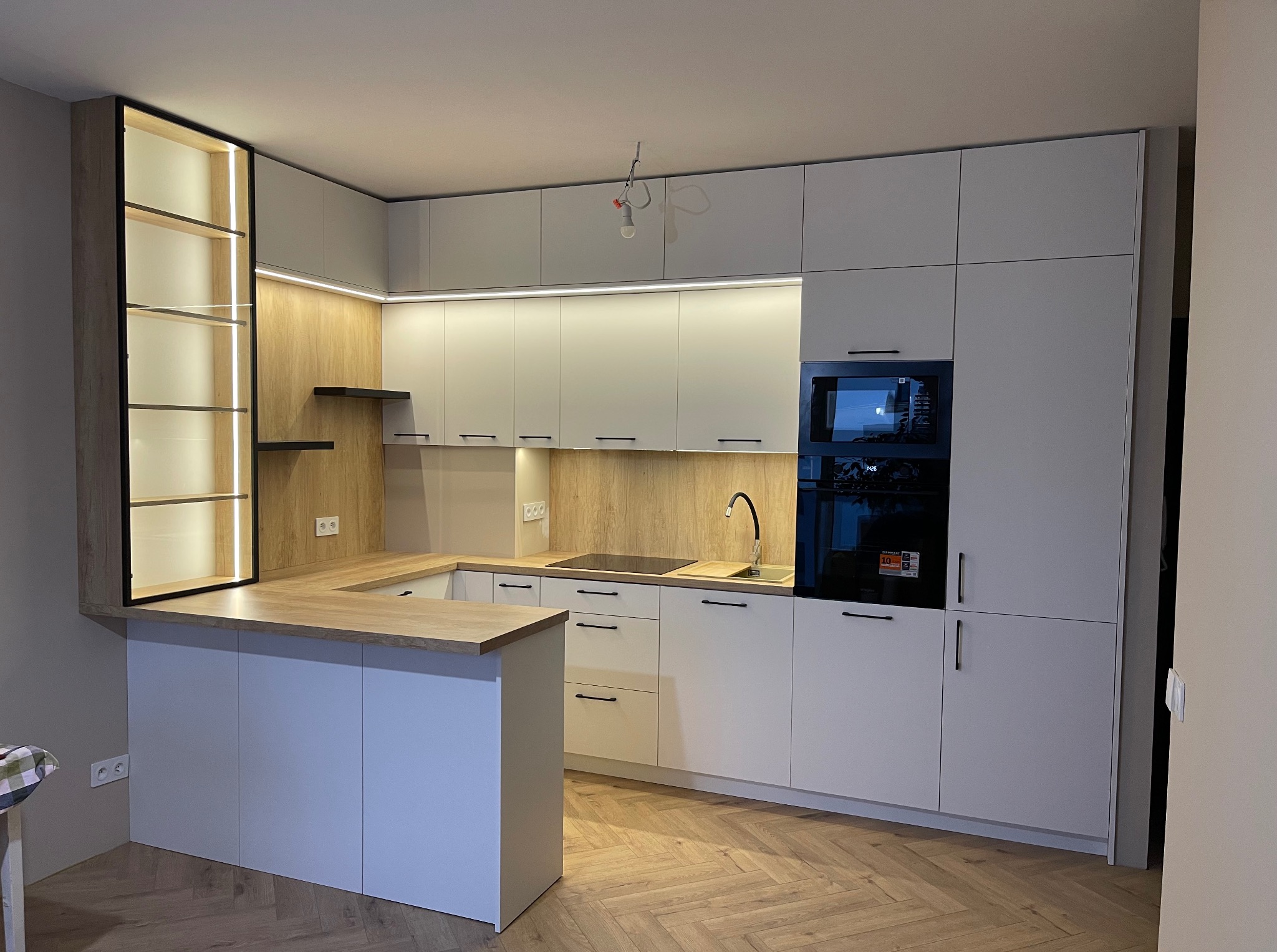 Nowoczesna kuchnia w bieli i drewnie z wyspą, oświetleniem LED i parkietem w jodełkę. Minimalistyczny design z czarnymi detalami i przeszkloną witryną.