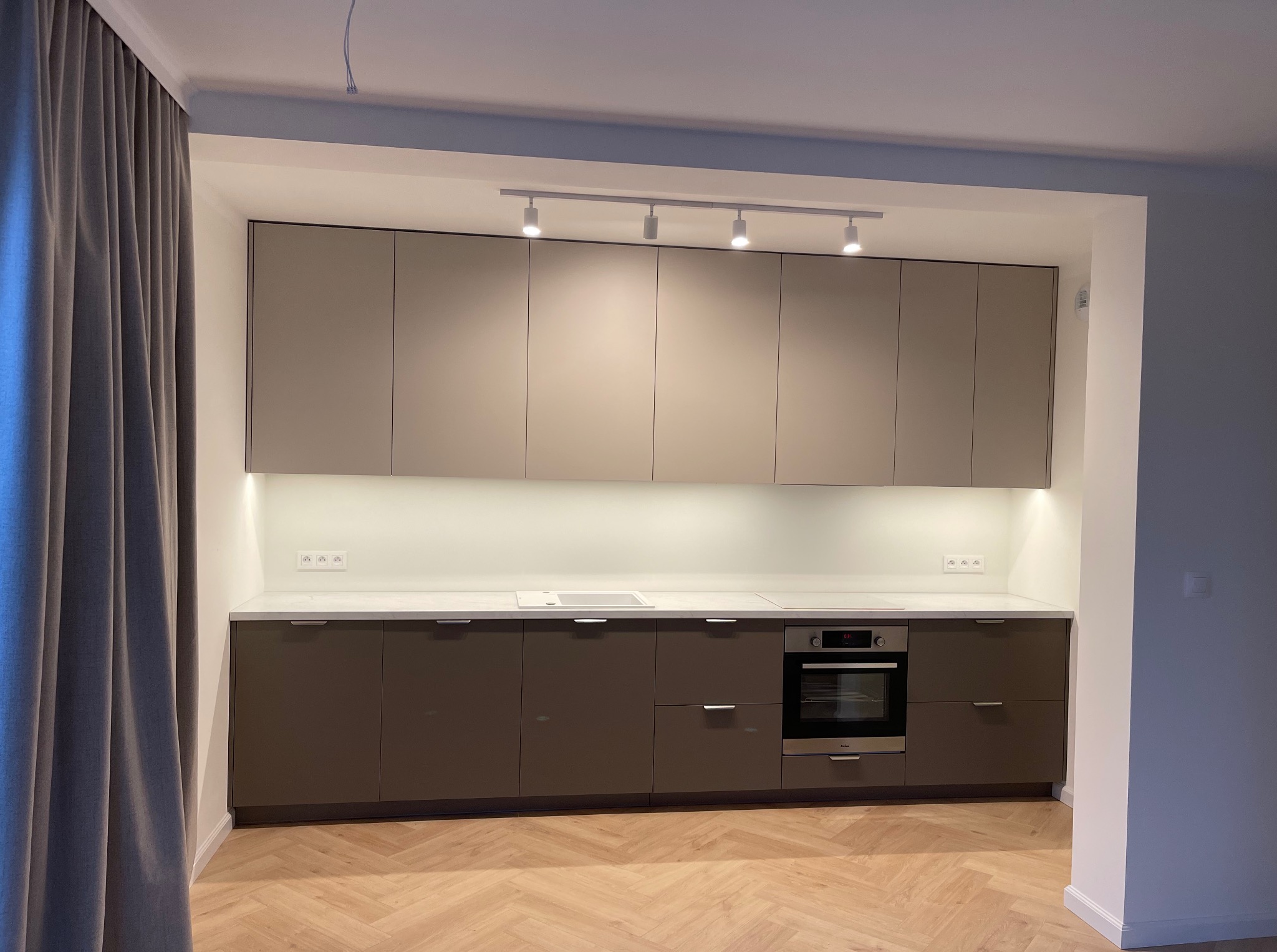 Nowoczesna kuchnia w odcieniach beżu i brązu z oświetleniem punktowym, jasnym blatem i drewnianą podłogą ułożoną w jodełkę. Widoczne zasłony po lewej stronie.