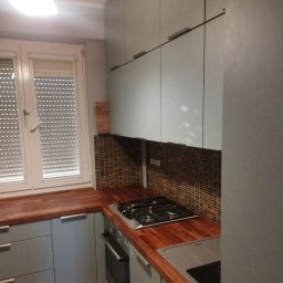 CORKY - Kuchnia z szarymi szafkami, drewnianym blatem i mozaiką nad blatem. Widoczne okno z żaluzjami i płyta grzewcza. Ujęcie z perspektywy.