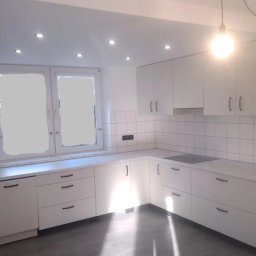 CORKY - Jasna kuchnia z białymi frontami i ciemnymi uchwytami, oświetlona punktowo i lampą wiszącą. Na ścianie białe płytki. Widoczne okno i szara podłoga.