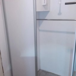 CORKY - Biała szafa wnękowa z przesuwnymi drzwiami i chromowanym drążkiem na ubrania. Widoczne fragmenty podłogi z jasnych płytek. Minimalistyczny design.