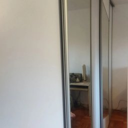 CORKY - Biała szafa wnękowa z przesuwnymi drzwiami i lustrem, odbijającym fragment pokoju z biurkiem i parkietem. Minimalistyczny design, widok z boku.