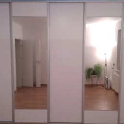 CORKY - Biała szafa wnękowa z przesuwanymi drzwiami i lustrami, odbijającymi wnętrze pokoju z drewnianą podłogą, rośliną doniczkową i lampą podłogową.