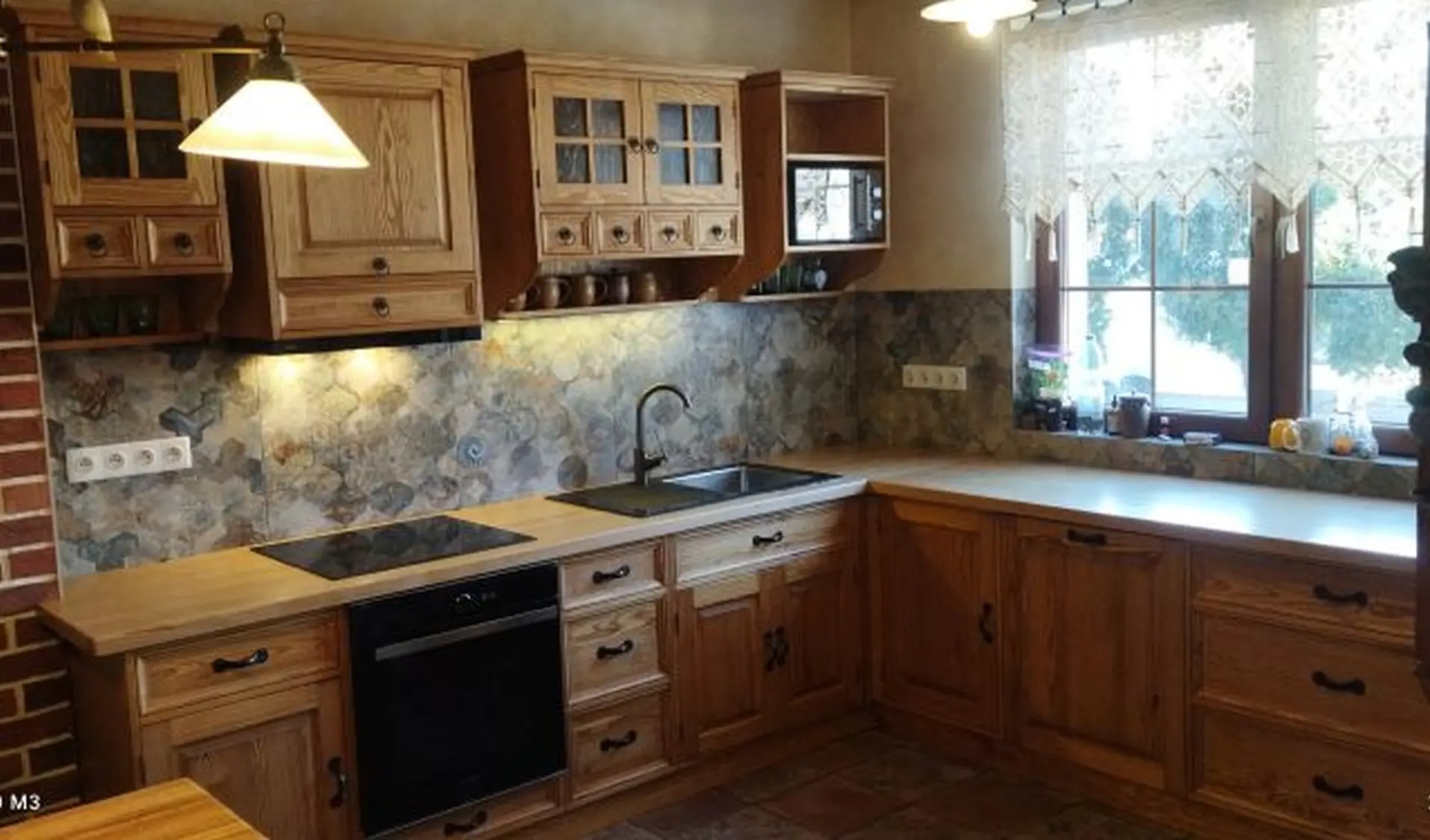 Rustykalna kuchnia z drewnianymi szafkami i blatem, okapem nad kuchenką, wzorzystą ścianą nad blatem i oknem z koronkową firanką.