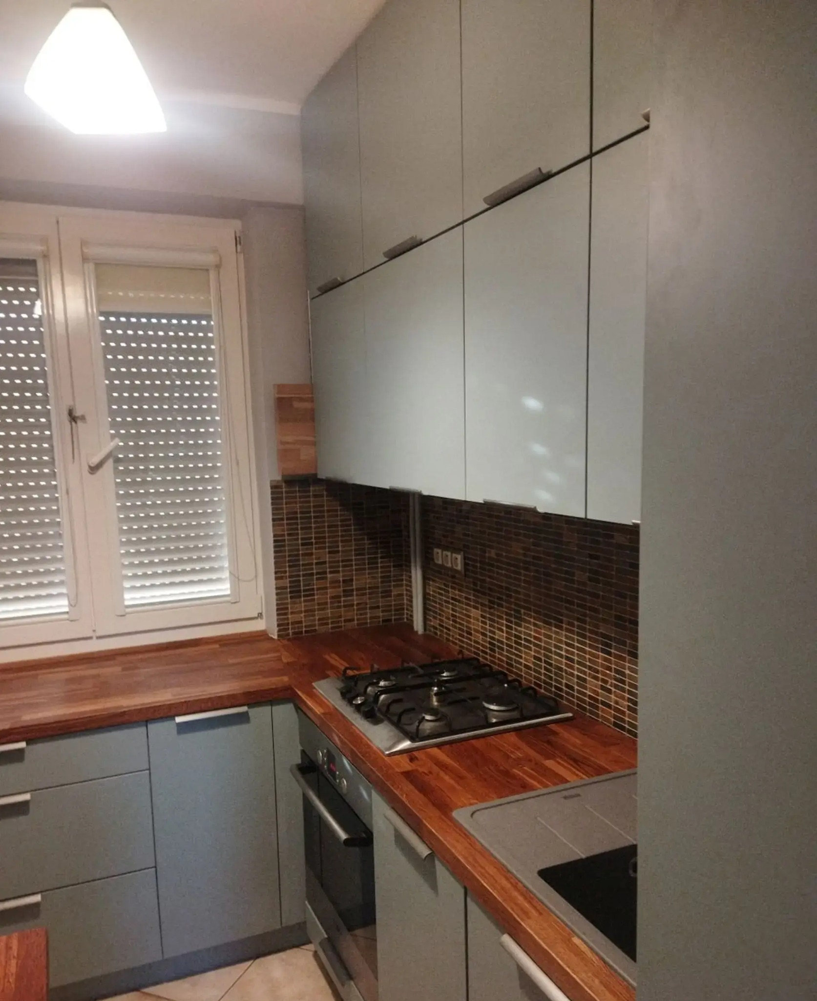 Kuchnia z szarymi szafkami, drewnianym blatem i mozaiką nad blatem. Widoczne okno z żaluzjami i płyta grzewcza. Ujęcie z perspektywy.