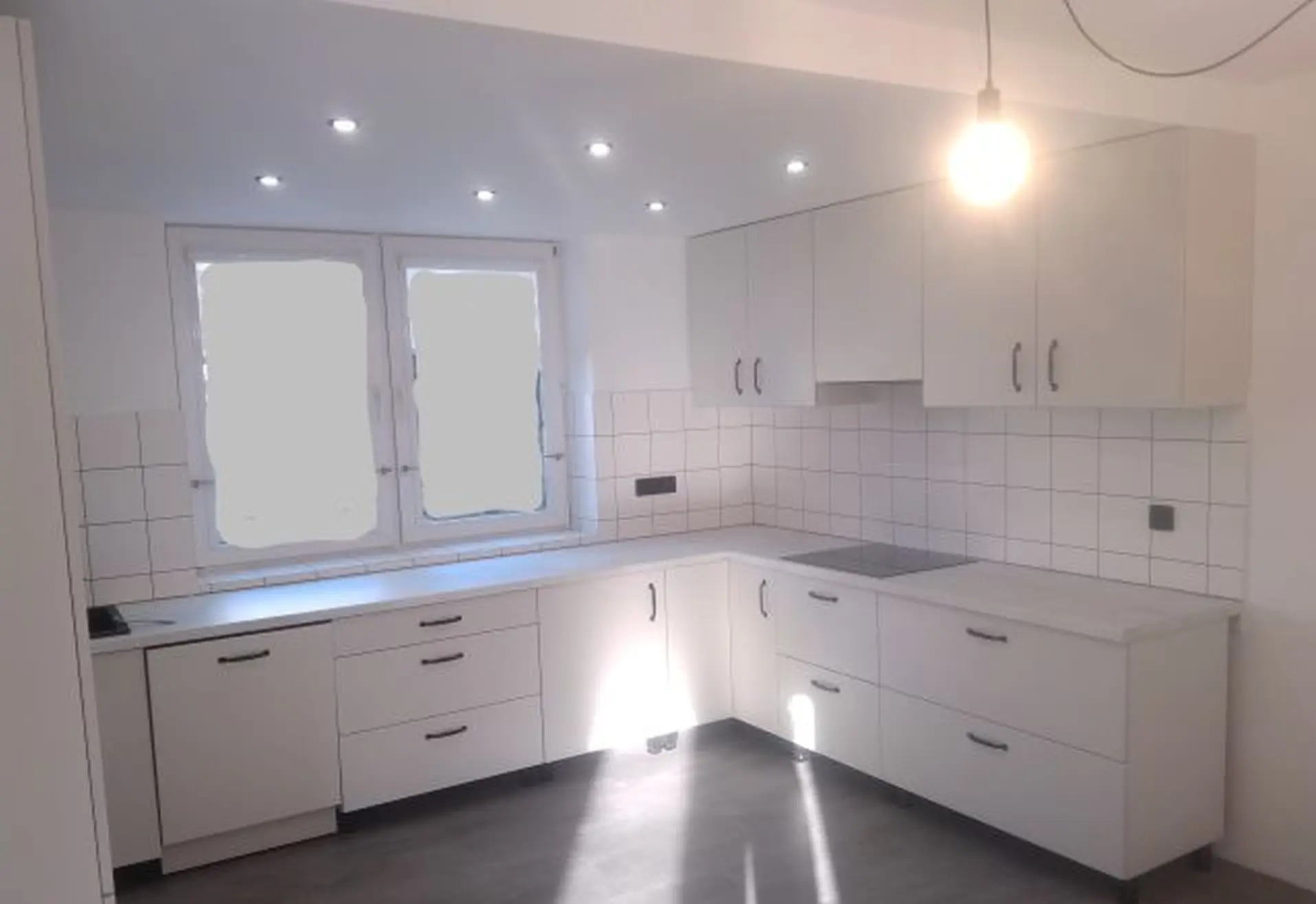 Jasna kuchnia z białymi frontami i ciemnymi uchwytami, oświetlona punktowo i lampą wiszącą. Na ścianie białe płytki. Widoczne okno i szara podłoga.