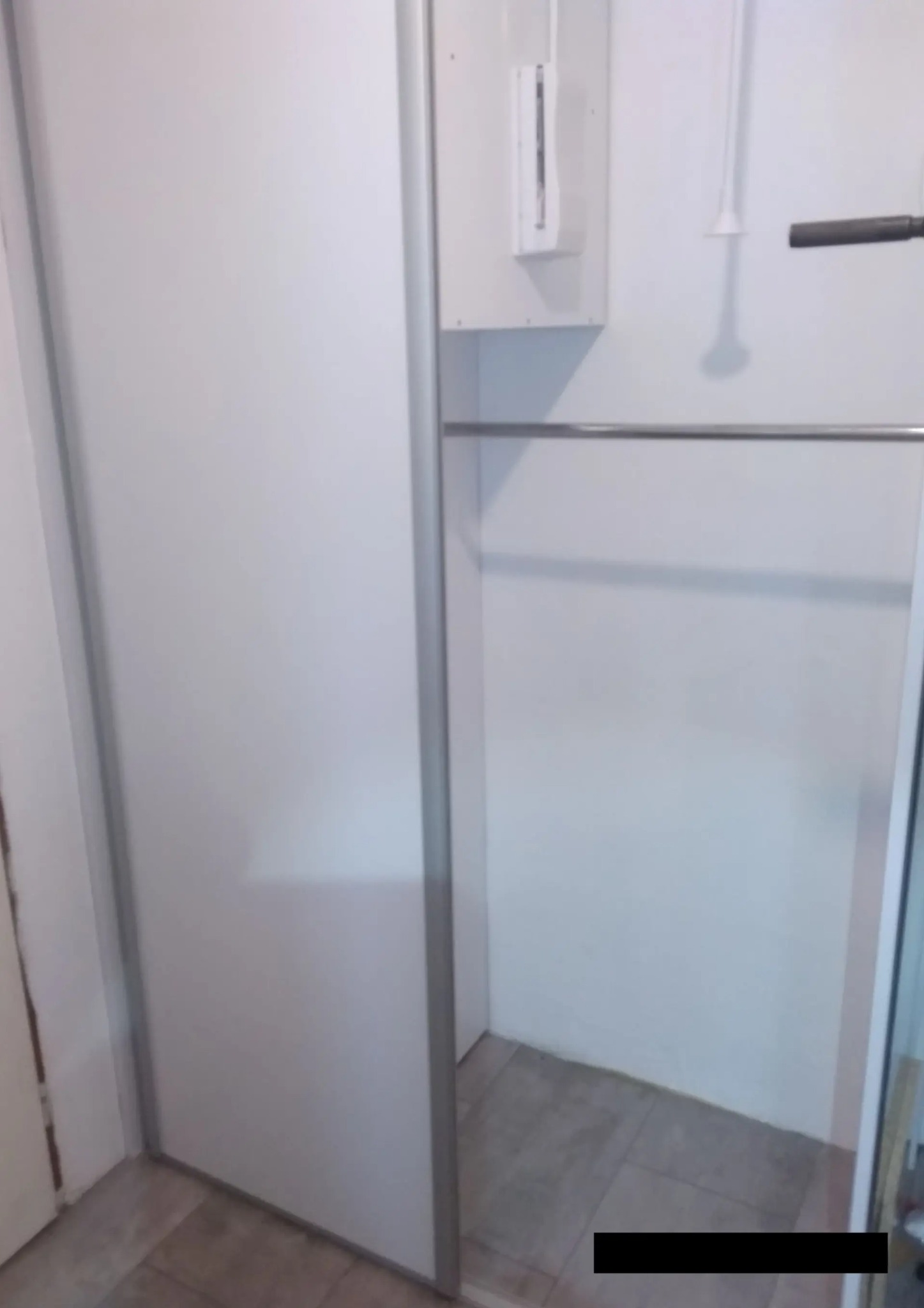 Biała szafa wnękowa z przesuwnymi drzwiami i chromowanym drążkiem na ubrania. Widoczne fragmenty podłogi z jasnych płytek. Minimalistyczny design.