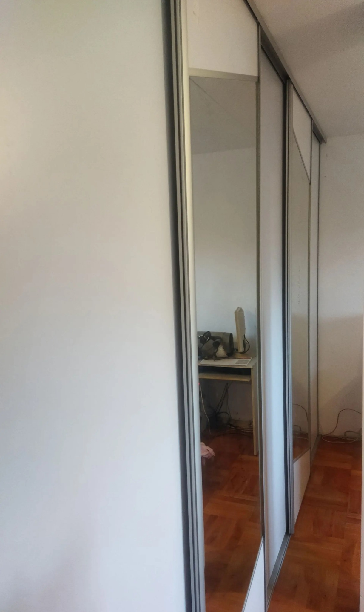 Biała szafa wnękowa z przesuwnymi drzwiami i lustrem, odbijającym fragment pokoju z biurkiem i parkietem. Minimalistyczny design, widok z boku.