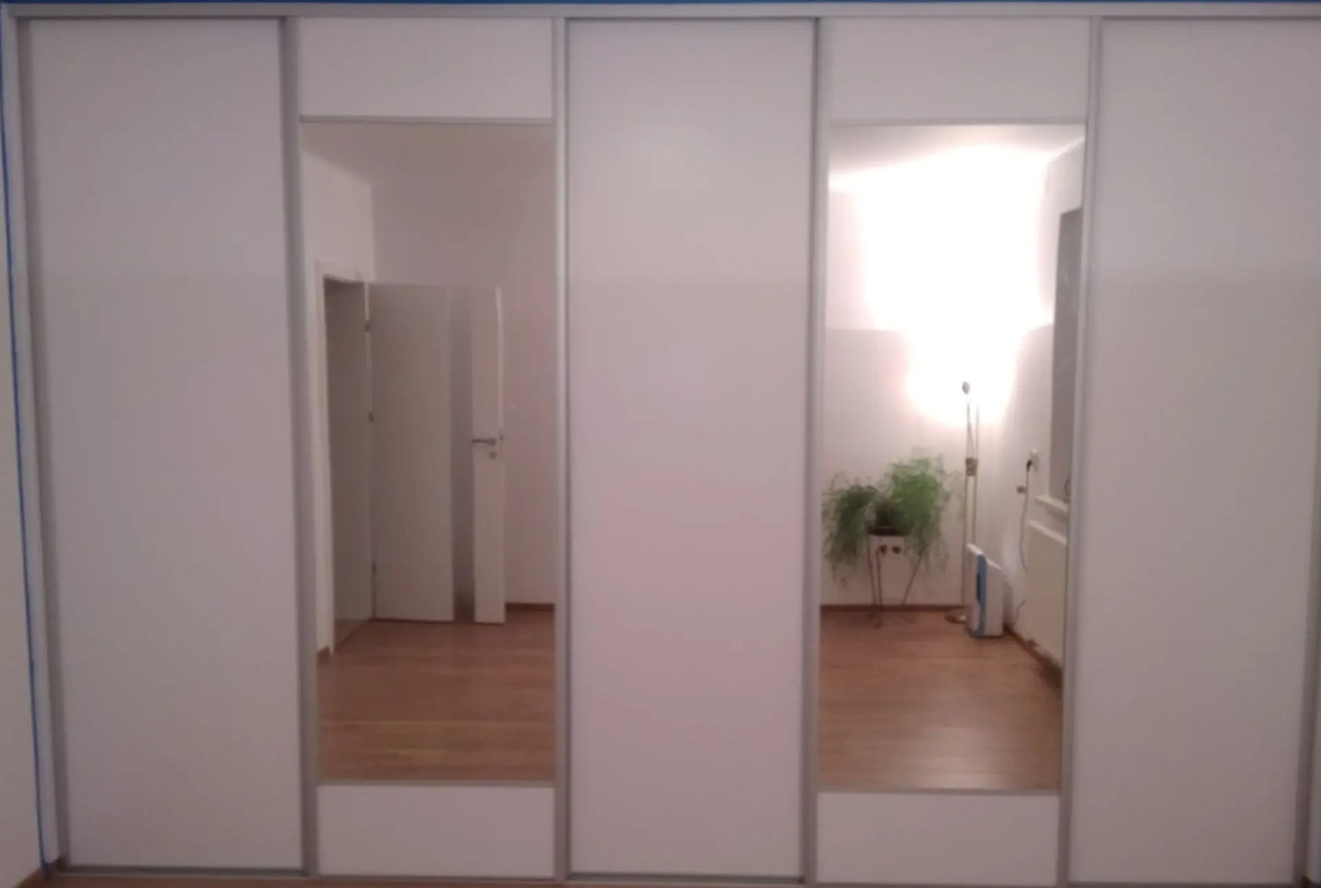 Biała szafa wnękowa z przesuwanymi drzwiami i lustrami, odbijającymi wnętrze pokoju z drewnianą podłogą, rośliną doniczkową i lampą podłogową.