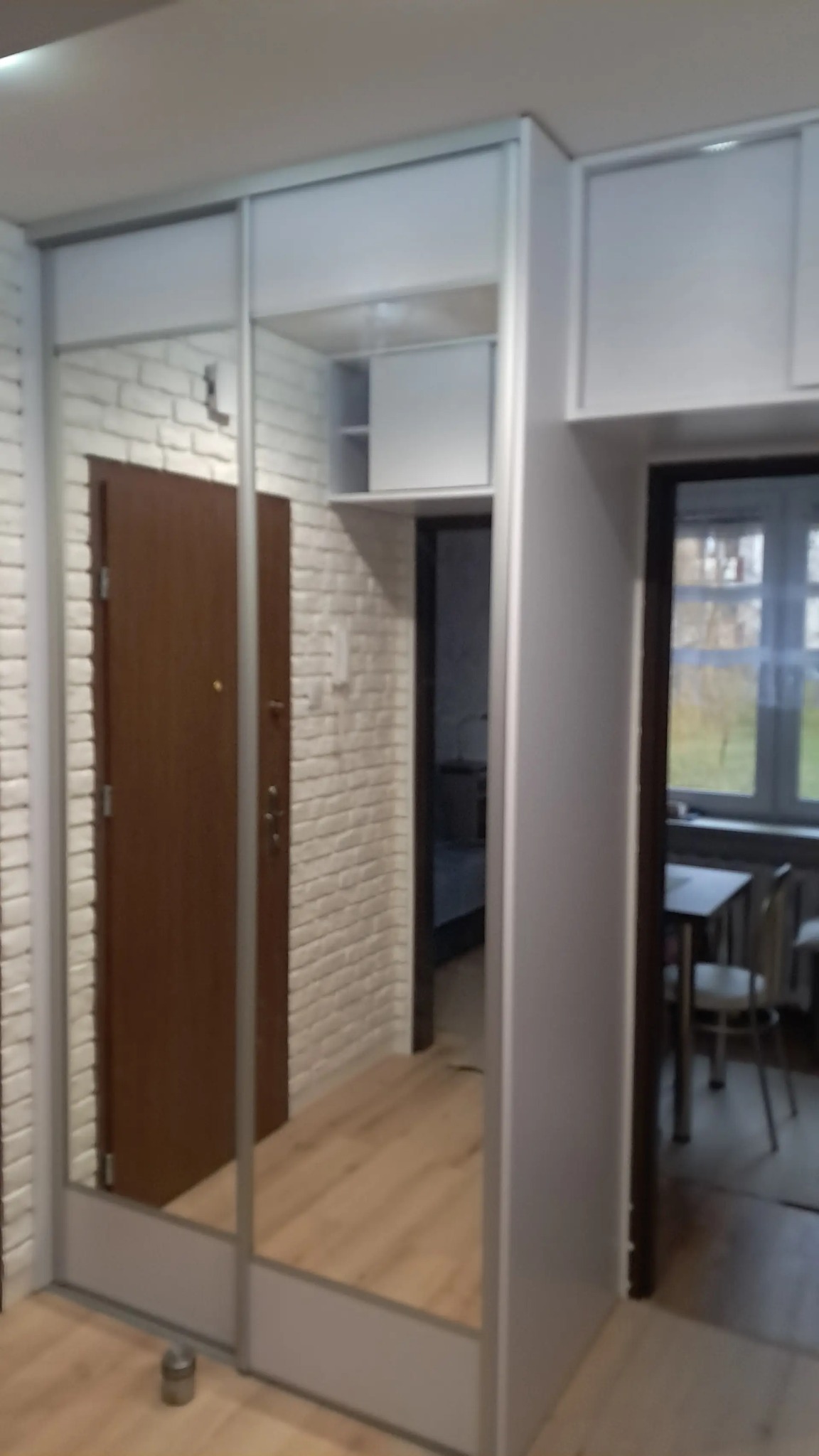 Biała szafa wnękowa z przesuwnymi lustrzanymi drzwiami, odbijająca brązowe drzwi wejściowe i białą ścianę z cegły w mieszkaniu.