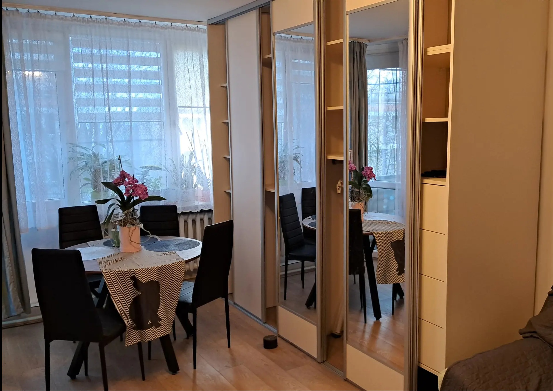 Jasna szafa przesuwna z lustrami w pokoju, odbijająca stół z kwiatami i krzesłami. Widoczny fragment łóżka i okno z firankami.