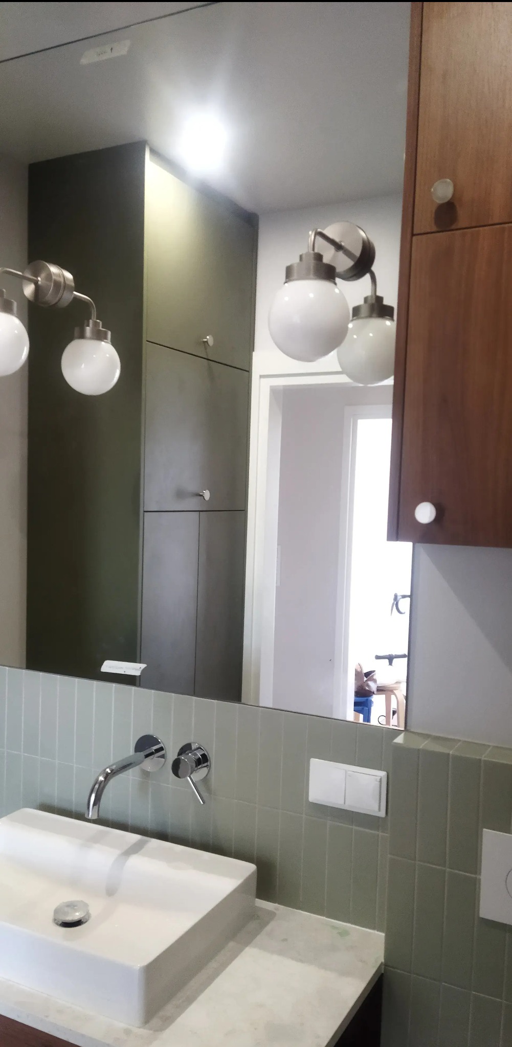 Nowoczesna łazienka z szafkami na wymiar w odcieniach brązu i zieleni, odbijającymi się w lustrze nad białą umywalką i baterią.