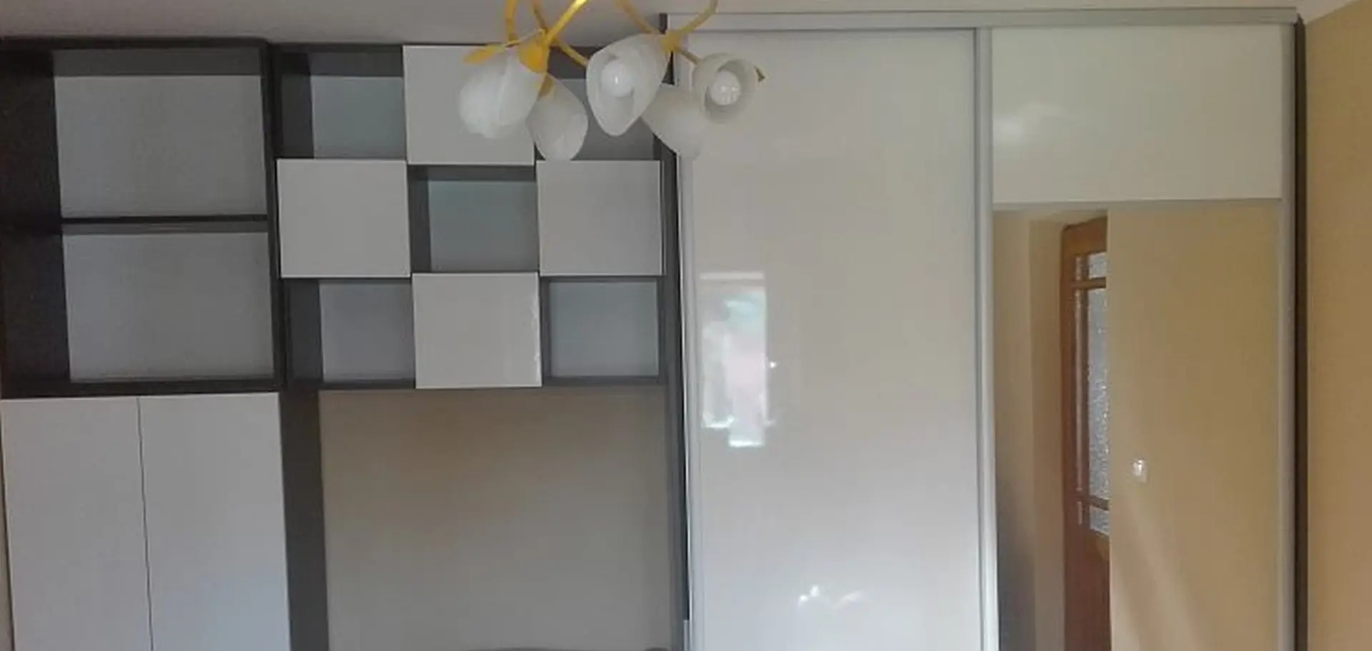 Biała szafa przesuwna z lustrem i regał z kwadratowymi półkami w odcieniach bieli i szarości, oświetlona lampą. Widok na pokój i drzwi.