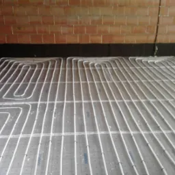 Białe rury ogrzewania podłogowego zamontowane na folii z rastrem, widoczne fragmenty ściany z cegły i izolacji. Instalacja przed wylaniem posadzki.