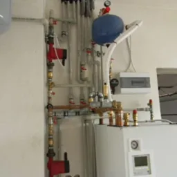 Złożona instalacja grzewcza z licznymi rurami, zaworami i pompami, zamontowana na ścianie, z widocznym zbiornikiem wyrównawczym i panelem sterowania.