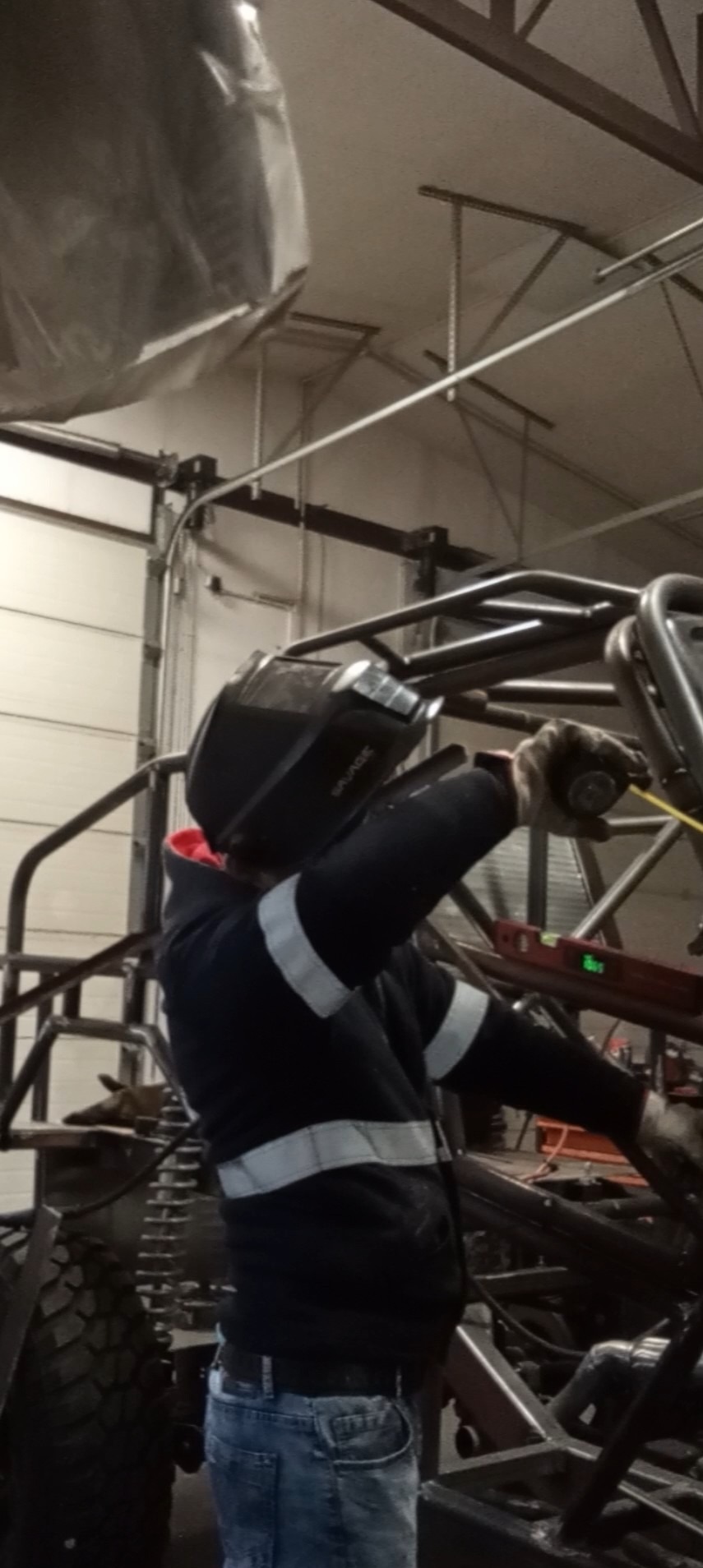 Pracownik w masce spawalniczej, w trakcie budowy metalowej konstrukcji pojazdu w warsztacie. Widoczne spawane rury, poziomica i opona.