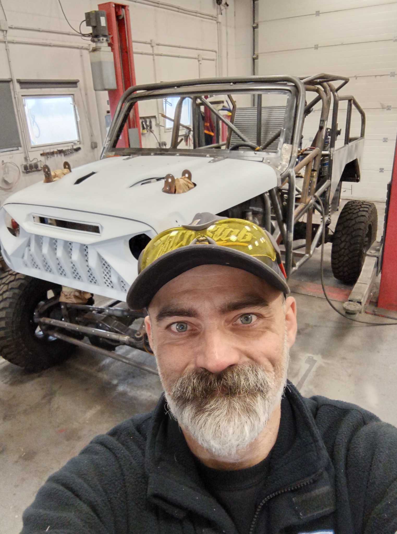 Selfie spawacza w okularach ochronnych na tle konstrukcji rurowej pojazdu terenowego na podnośniku warsztatowym. Widoczna spawana rama i elementy karoserii.