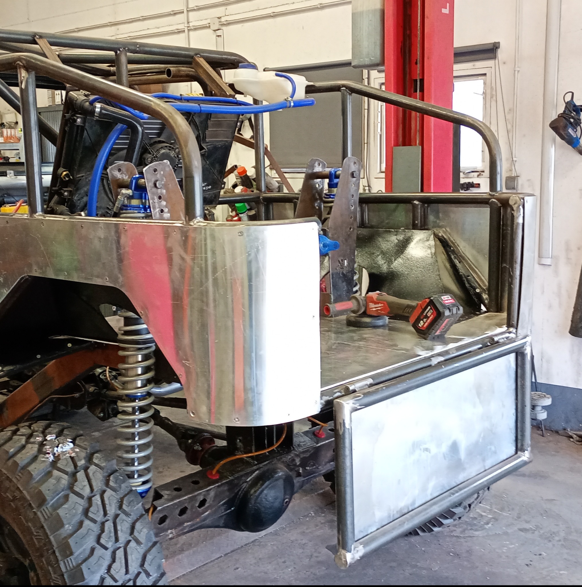 Rama pojazdu terenowego w trakcie budowy. Widoczne spawane elementy, amortyzatory, koło i narzędzia warsztatowe, w tym szlifierka kątowa.