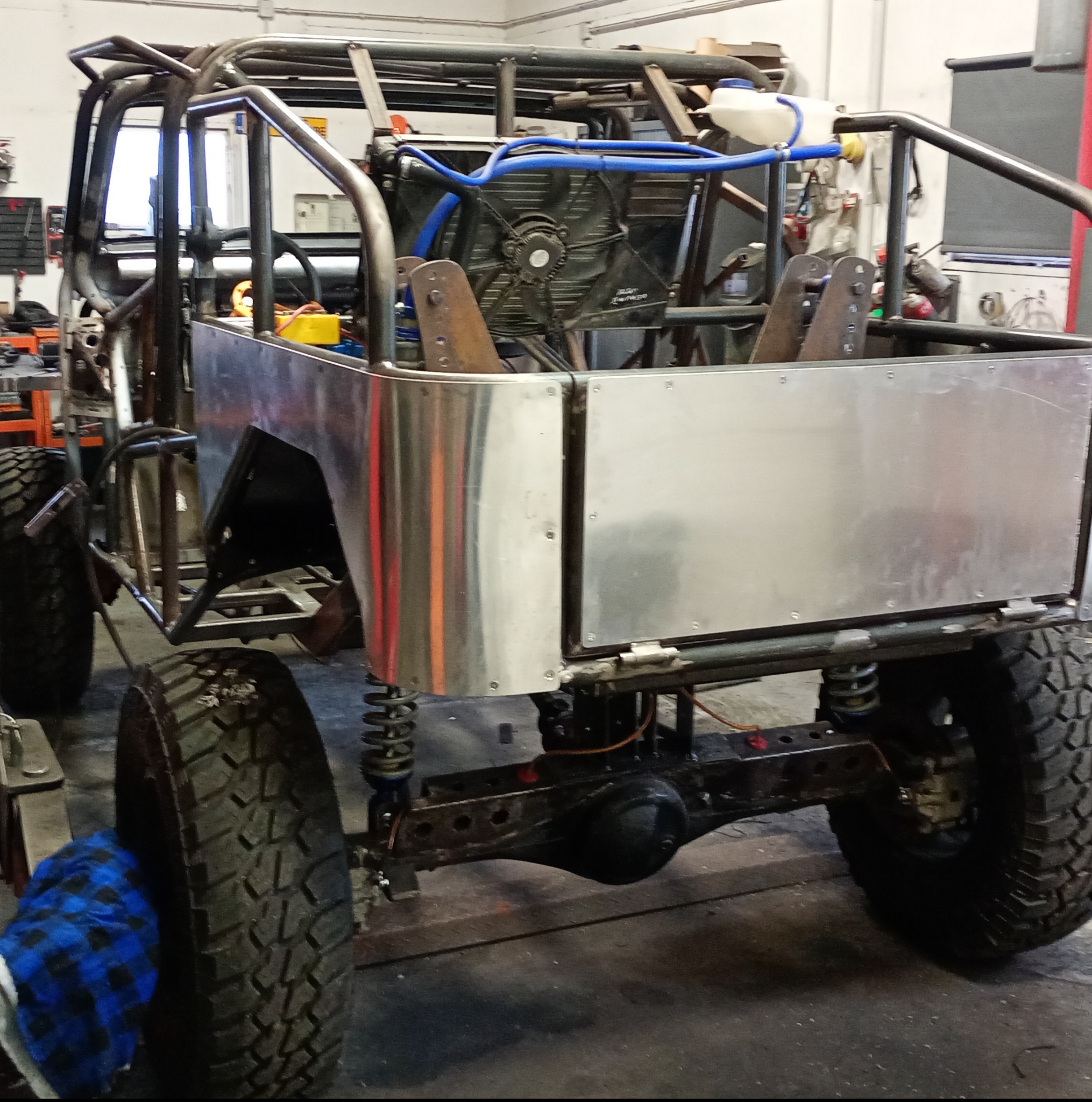 Rama pojazdu terenowego w trakcie budowy. Widoczne metalowe rury, części karoserii z blachy, opony terenowe i elementy zawieszenia. Warsztatowe otoczenie.