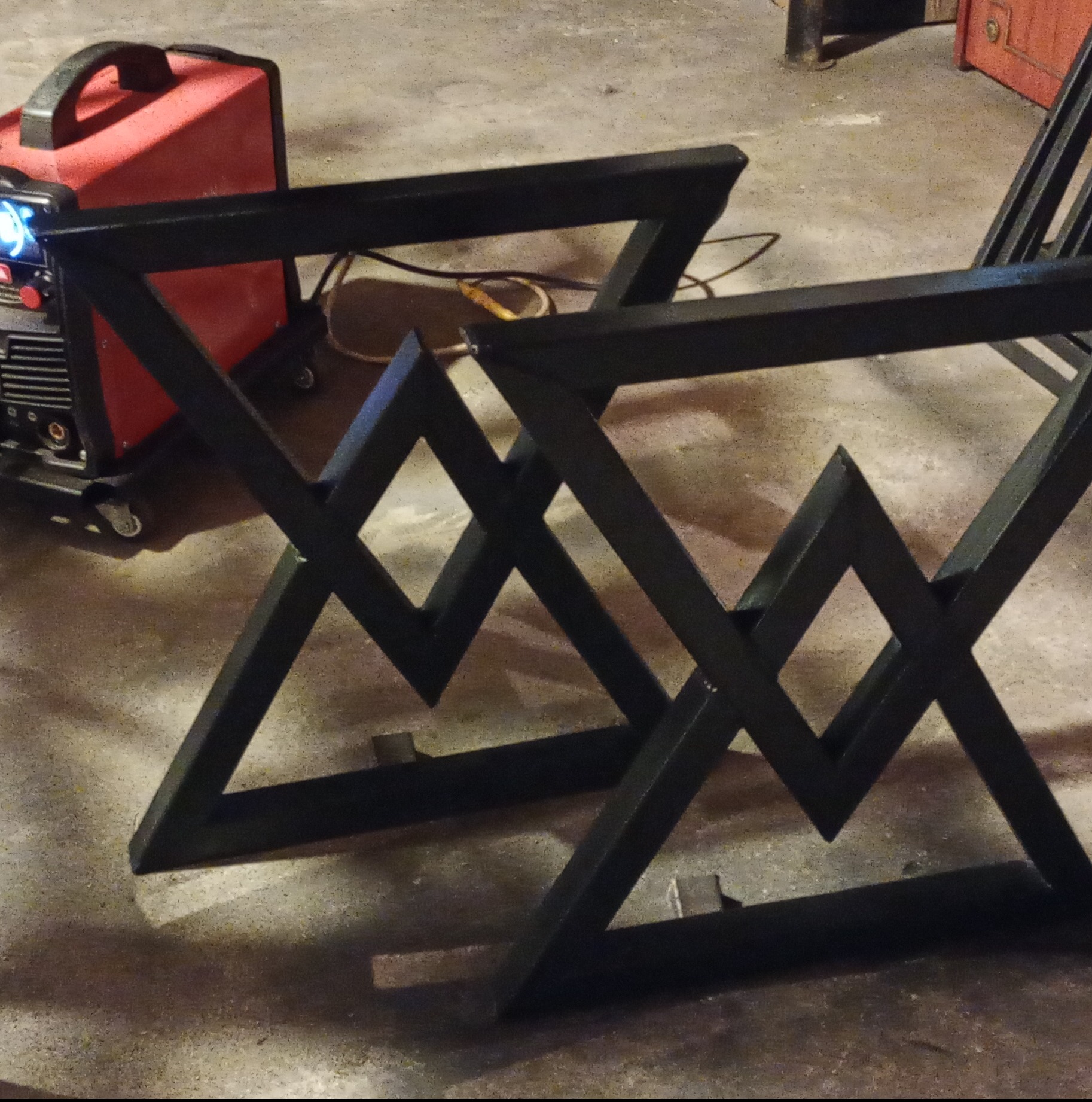 Dwie czarne, spawane podstawy stołu o geometrycznym wzorze, ustawione na szarym betonie, obok czerwonej spawarki z widocznymi kablami.