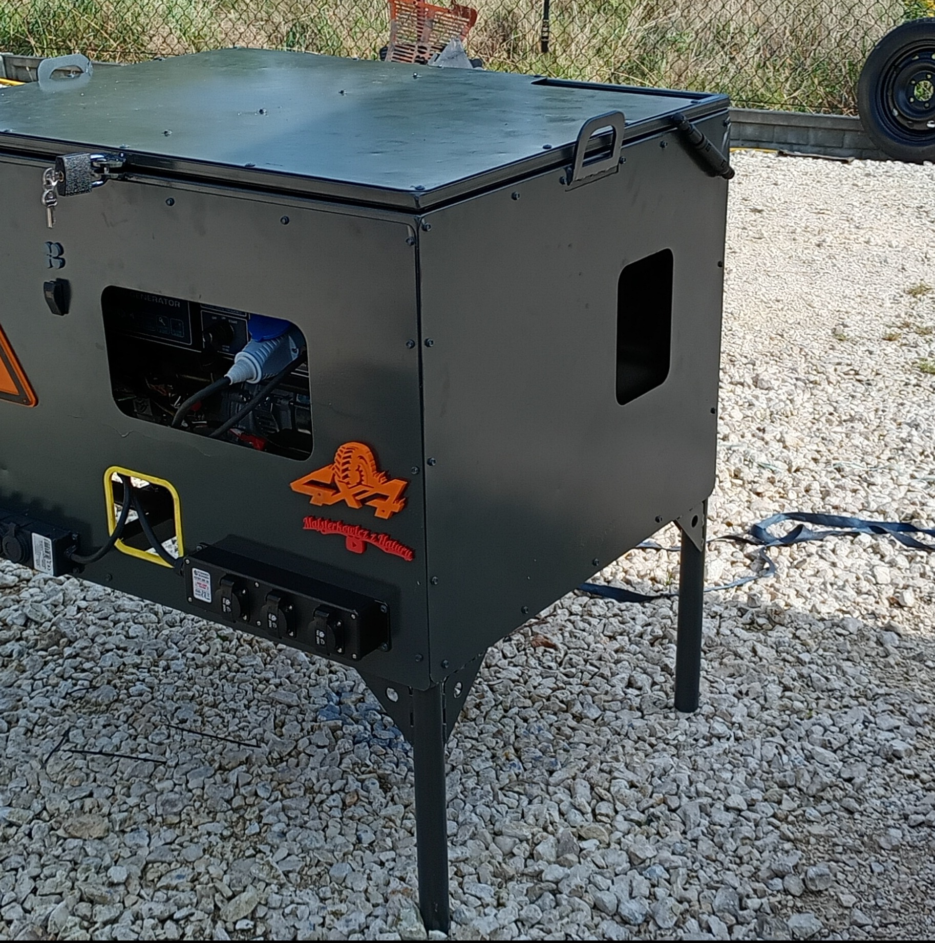 Czarna, metalowa skrzynia z generatorem i gniazdami, stojąca na czterech nogach. Logo 4x4. Widoczne detale spawania i mocowania. Realizacja z zakresu obróbki metali.