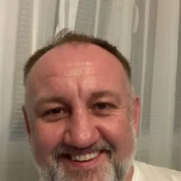 Uśmiechnięty mężczyzna z brodą na tle białej zasłony, portret w naturalnym świetle.