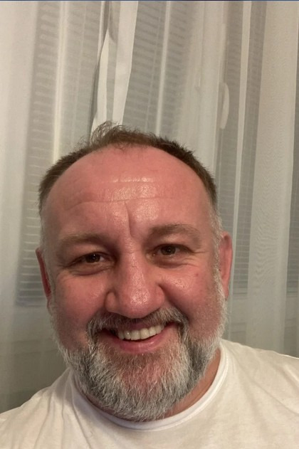 Uśmiechnięty mężczyzna z brodą na tle białej zasłony, portret w naturalnym świetle.