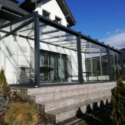 Szklana zabudowa tarasu z czarną ramą, widok z zewnątrz. Stolik i krzesła wewnątrz. Schody z płytek prowadzą na taras. Błękitne niebo w tle.