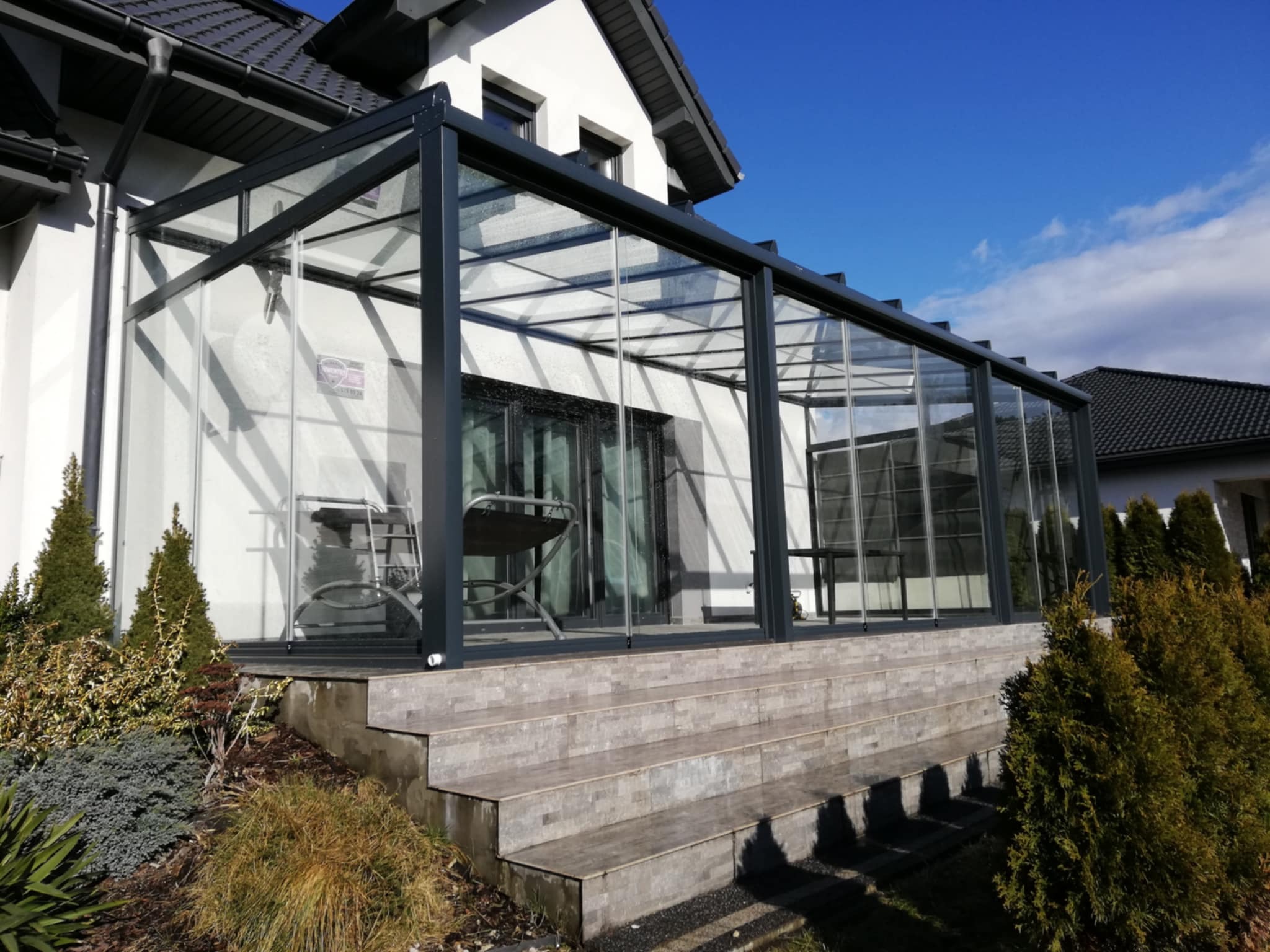 Szklana zabudowa tarasu z czarną ramą, widok z zewnątrz. Stolik i krzesła wewnątrz. Schody z płytek prowadzą na taras. Błękitne niebo w tle.