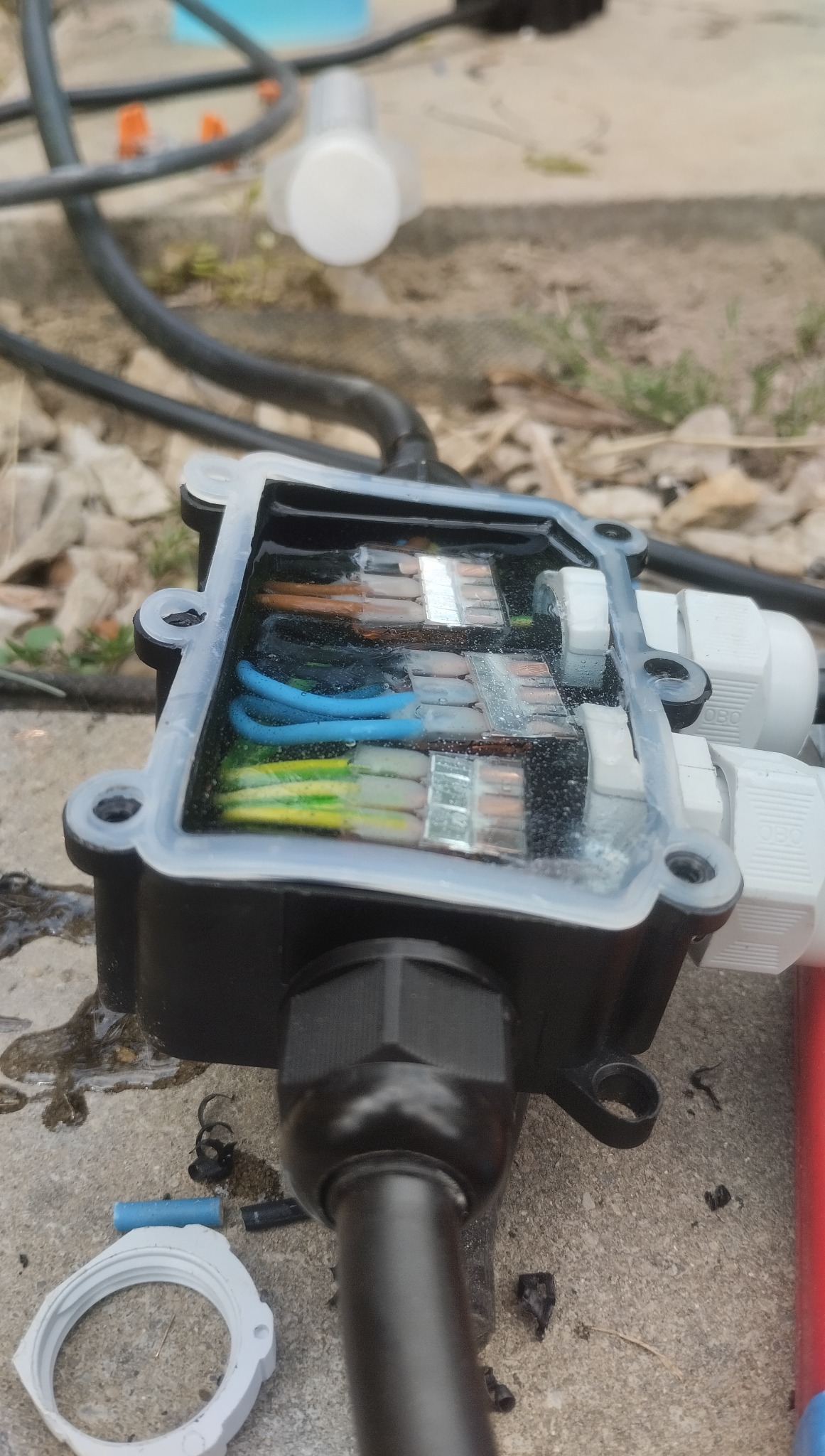 Otwarta, czarna puszka instalacyjna z widocznymi wewnątrz złączkami WAGO i przewodami w kolorach: brązowym, niebieskim i zielono-żółtym. Na zewnątrz fragment kabla i elementy montażowe.