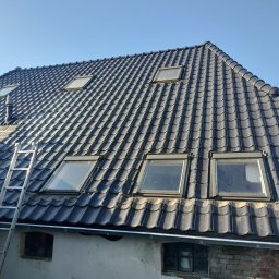 Dariusz Partyczny - Czarny dach z dachówki ceramicznej z oknami połaciowymi, widziany pod kątem. Aluminiowa drabina oparta o ścianę. Błękitne niebo w tle.