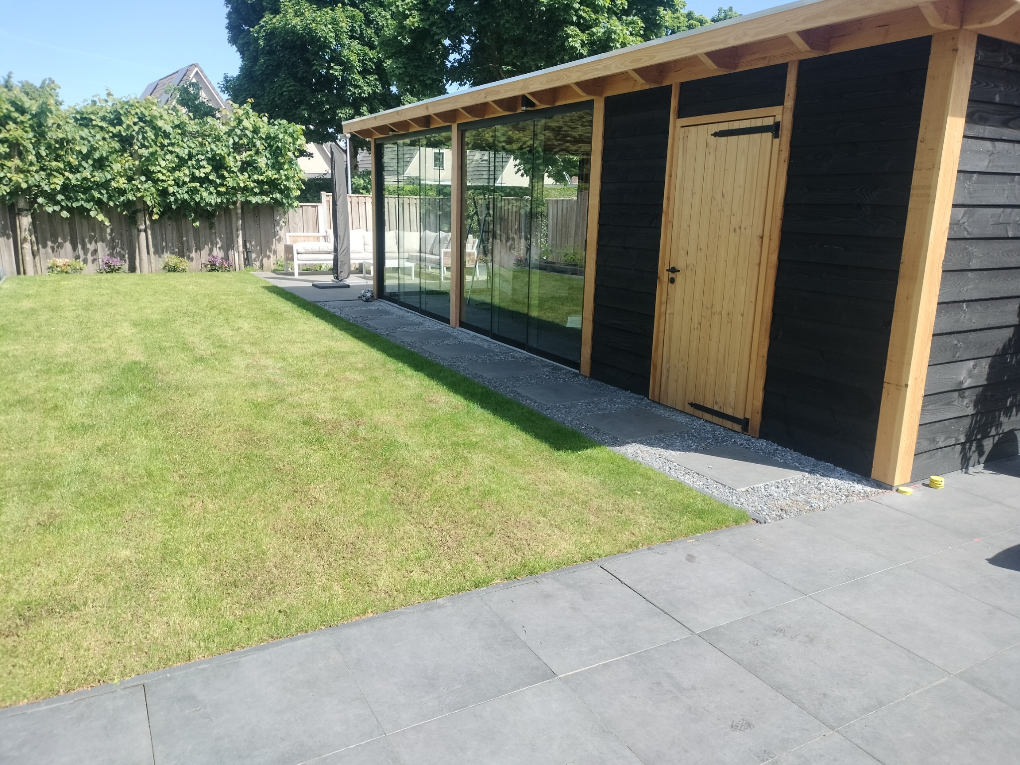 Nowoczesna altana z drewnianą konstrukcją i szklanymi ścianami we Wrocławiu. Czarne deski elewacyjne kontrastują z jasnym drewnem drzwi i ramy. Zadbane otoczenie z trawnikiem.