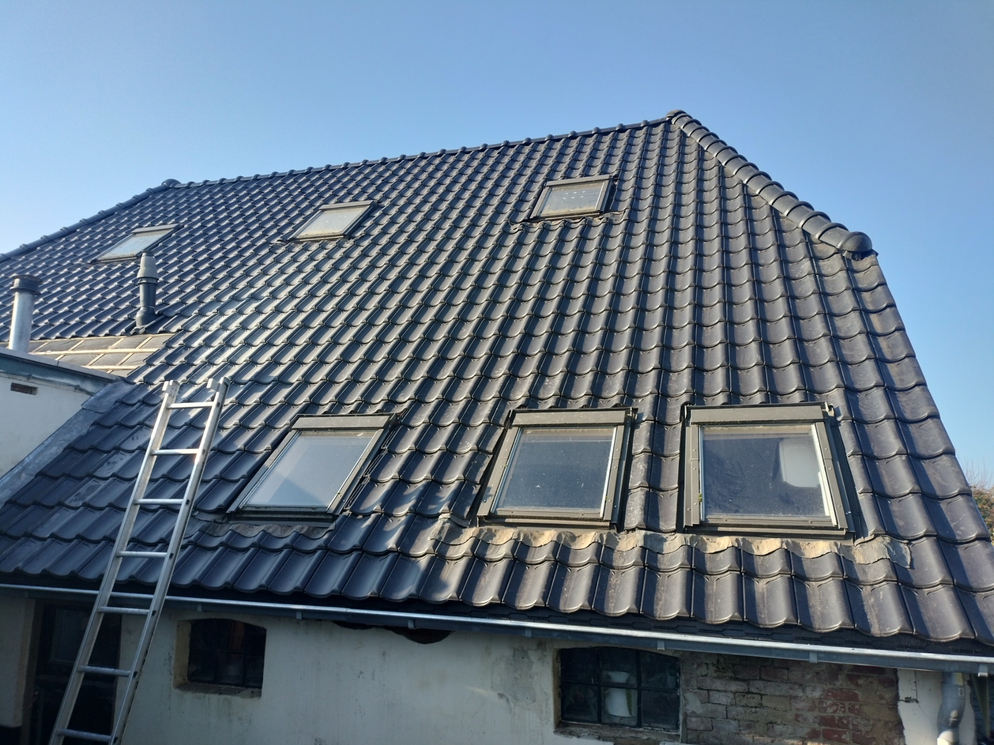Czarny dach z dachówki ceramicznej z oknami połaciowymi, widziany pod kątem. Aluminiowa drabina oparta o ścianę. Błękitne niebo w tle.