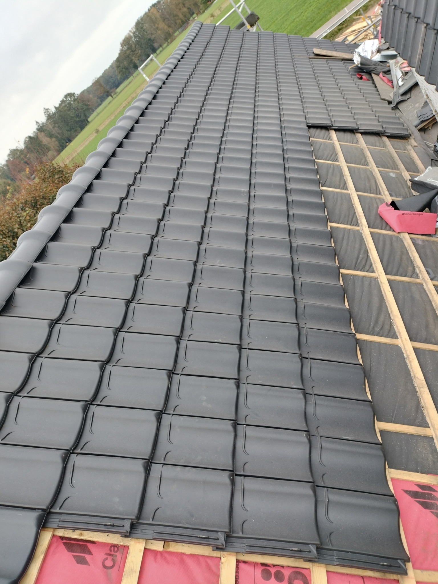 Częściowo pokryty dach czarną dachówką ceramiczną. Widoczne łaty i membrana dachowa. W tle zielony krajobraz.