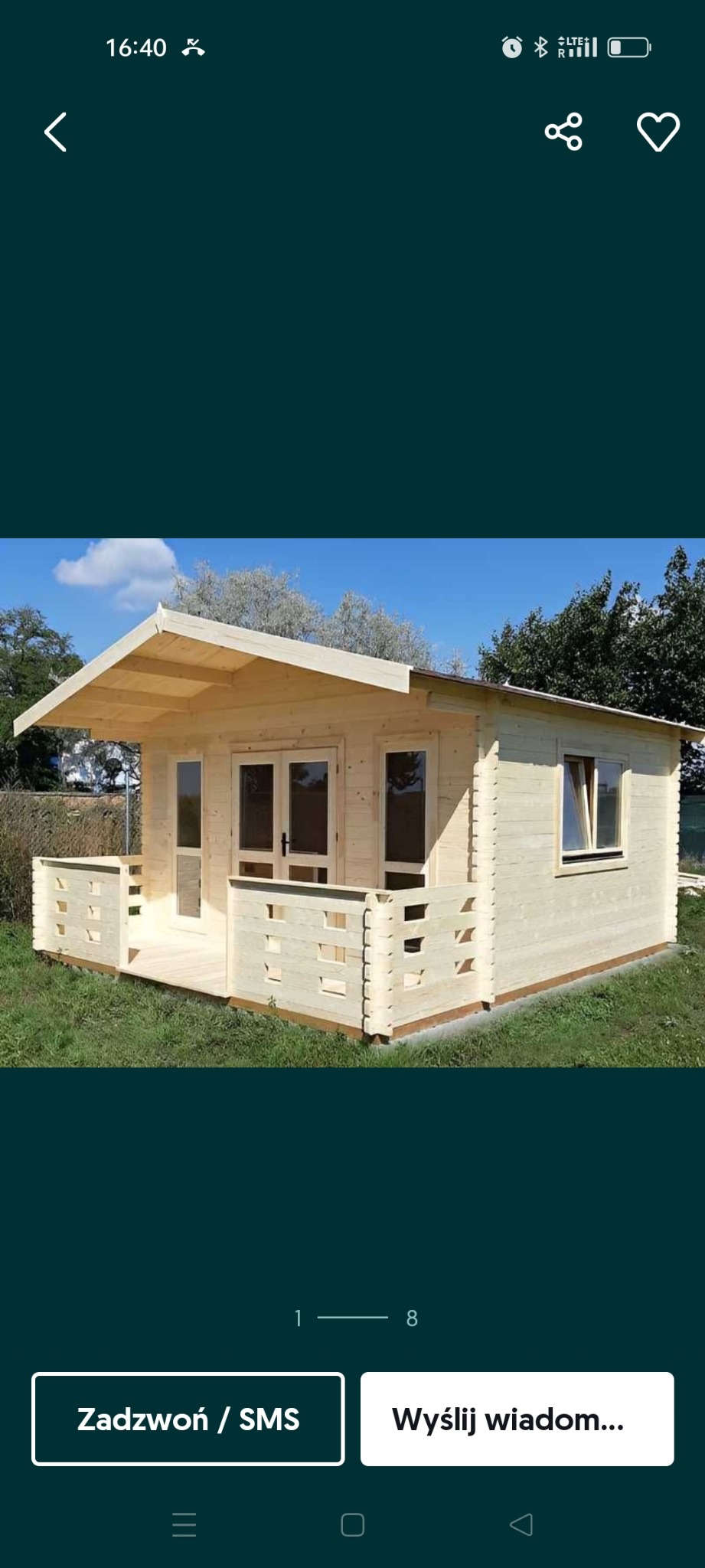 Drewniany domek letniskowy z werandą, jasne drewno, dwuskrzydłowe drzwi z przeszkleniami, okno z okiennicą, trawnik, błękitne niebo w tle.