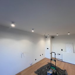 eFLOW - Wnętrze po malowaniu ścian na biało, widoczne nowe lampy sufitowe, zakryta podłoga i drabina malarska. Świeżo wykończone gniazdka elektryczne.
