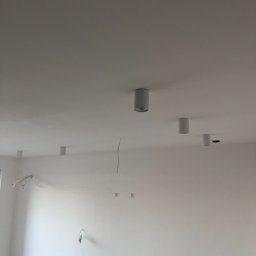 eFLOW - Białe, cylindryczne lampy sufitowe zamontowane w jasnym pomieszczeniu. Widoczne przewody i przygotowane punkty świetlne na ścianie. Minimalizm i surowość wnętrza.